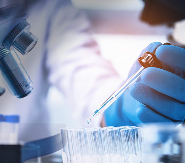 A gloved hand holds a pipette above multiple test tubes, with a microscope in the background.