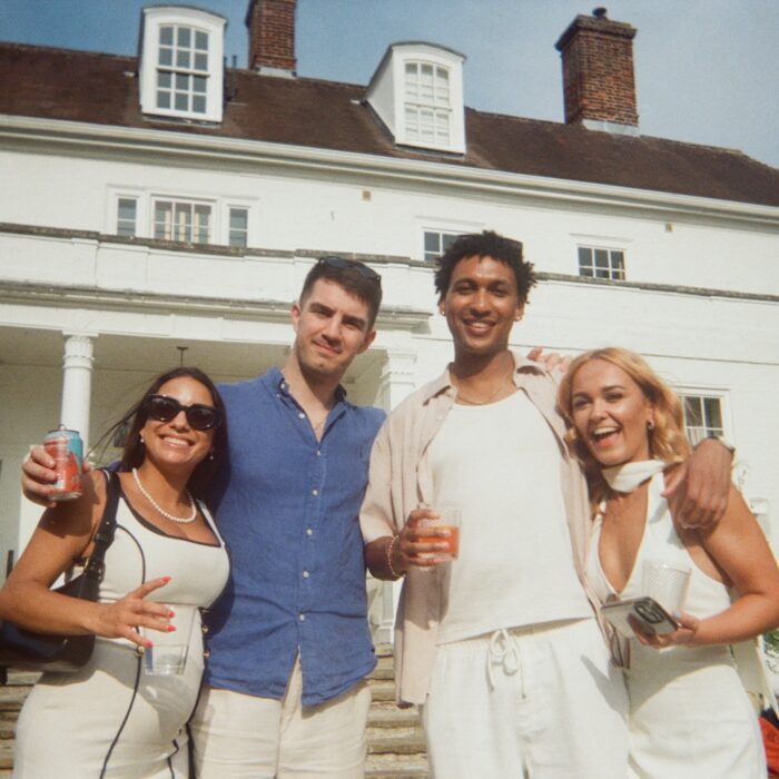 Four friends pose for a photo in front of a large white house, smiling and holding drinks.