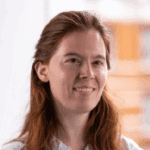 A woman with long red hair and a slight smile, wearing a light-colored shirt.