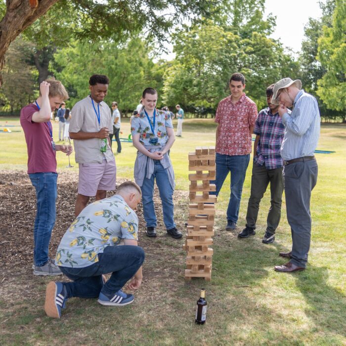 A group of six people gathers around a towering Jenga-like game, focused on the wooden blocks. One person kneels beside the structure while others stand in anticipation. They are dressed in casual summer attire, with some wearing colorful shirts. A drink is