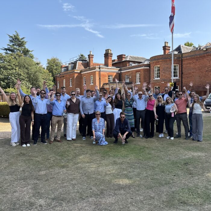 A large group of people standing outdoors, smiling and waving, with a historic building and a flag in the background.