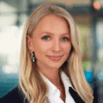 A professional woman with long blonde hair and earrings smiles warmly, set against a blurred office background.
