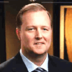 A smiling man in a suit, standing in a professional setting.