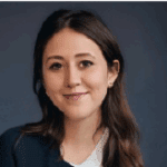 A woman smiling warmly, with long brown hair, wearing a dark blazer over a white top, against a dark blue background.
