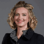 Smiling woman with curly blonde hair, wearing a dark blazer over a patterned shirt.
