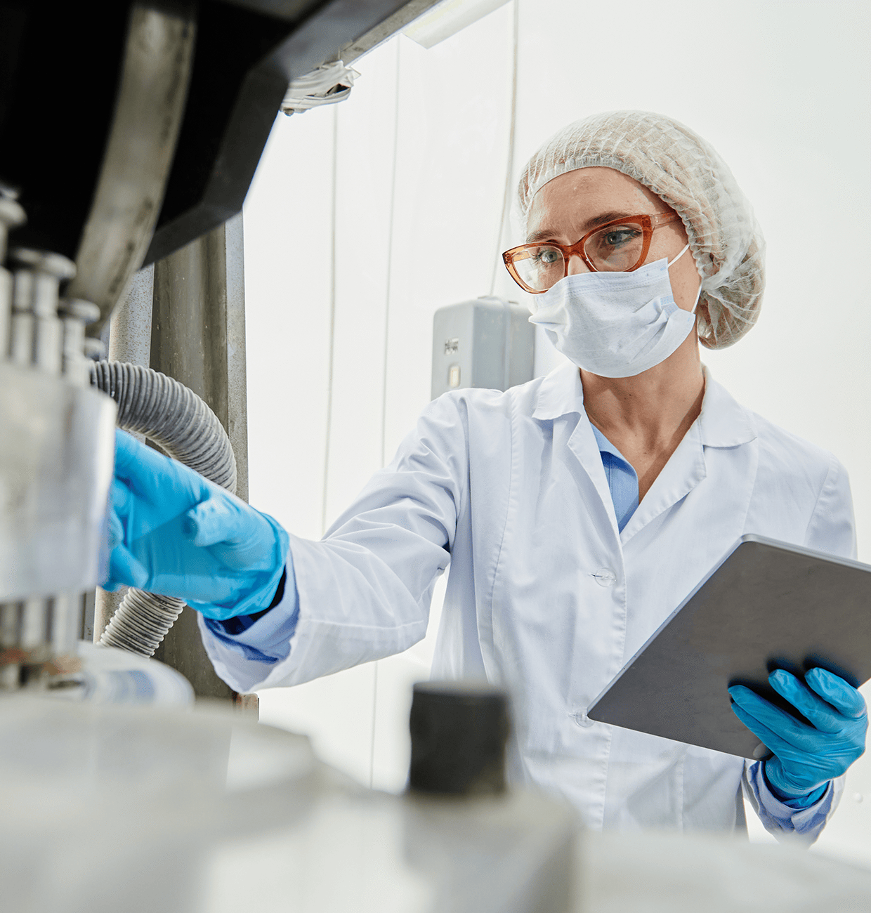 A laboratory technician in a white coat and gloves works with equipment while holding a tablet. They wear a hairnet and face mask, focusing on a machine.